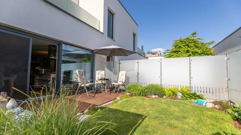 Moderne Terrasse mit Holzdeck, Gartenmöbeln und kleinem Rasenbereich vor einem weißen Gebäude.