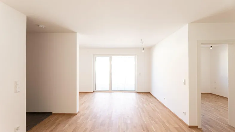 Helles, leeres Zimmer mit Holzboden und Zugang zu einem Balkon oder einer Terrasse.