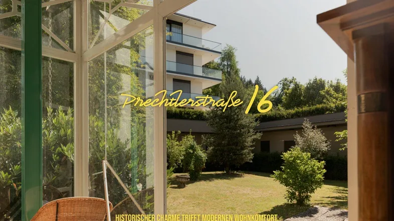 Blick aus einem verglasten Wintergarten mit Korbsessel auf einen gepflegten Garten und ein modernes Gebäude.