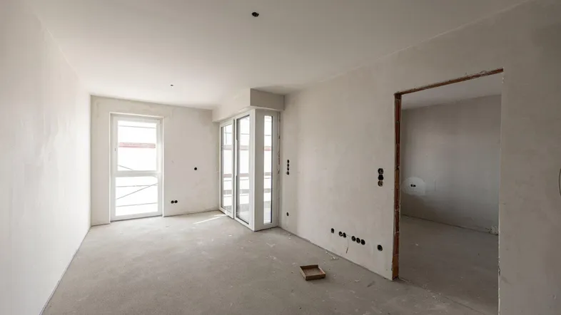 Unfertiger Innenraum mit Betonboden und großen Fenstern, bereit für den Ausbau.