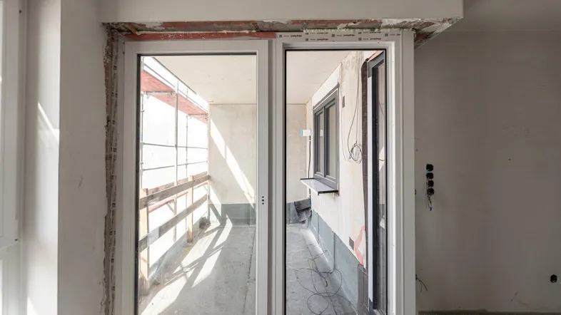 Unfertiger Innenraum mit großen Fenstern und Zugang zu einem zukünftigen Balkon.