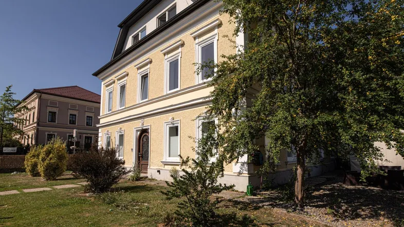 Historisches Gebäude mit gelber Fassade und gepflegtem Vorgarten unter blauem Himmel.