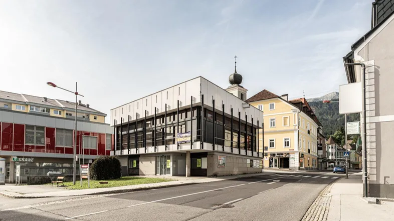 Modernes Geschäftsgebäude an einer belebten Straße mit umliegenden Häusern und Bergen im Hintergrund.