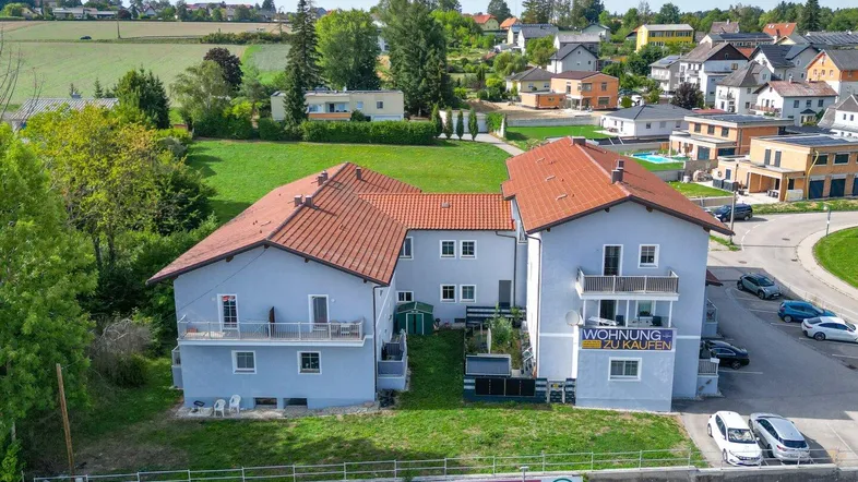 Luftaufnahme eines Mehrfamilienhauses mit hellgrauer Fassade, roten Ziegeldächern und umliegender Grünfläche.