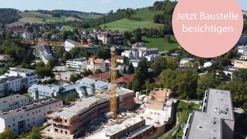 Luftaufnahme einer Baustelle mit neuen Gebäuden und einem Kran, umgeben von grünen Hügeln und Wohngebieten.