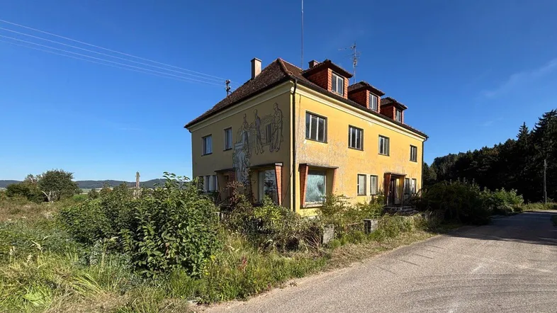 Ein älteres, gelb gestrichenes Gebäude mit rotem Ziegeldach und umliegender Natur unter blauem Himmel.