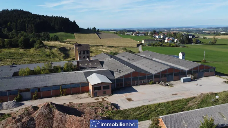 Luftaufnahme eines Industrieareals mit mehreren Hallen und einem Turm, umgeben von grünen Feldern und Wäldern.