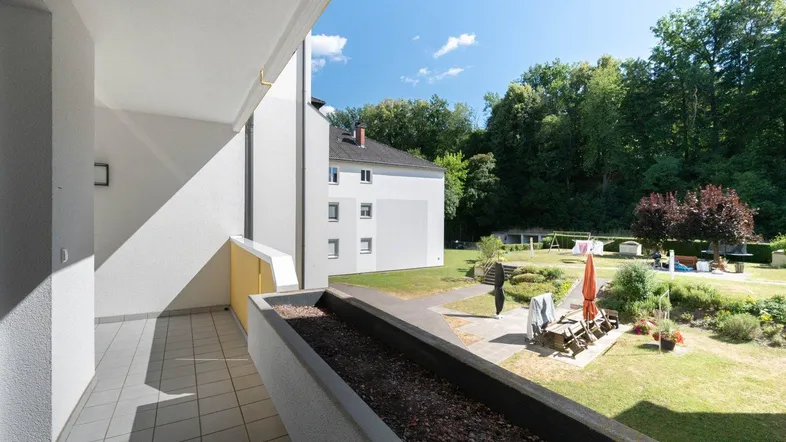 Heller Balkon mit Fliesenboden und Blick auf den gepflegten Gemeinschaftsgarten mit Spielplatz und Bäumen.