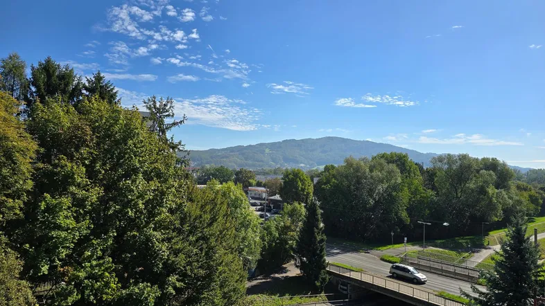 Weitläufiger Blick auf grüne Hügel und eine Straße mit Brücke unter blauem Himmel.