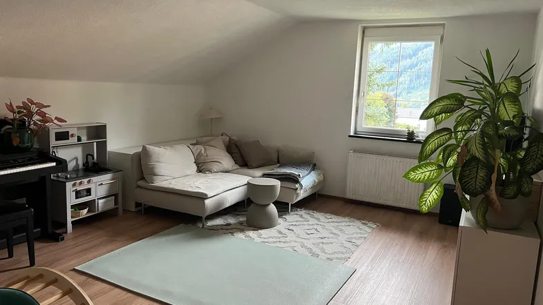 Helles Wohnzimmer mit Dachschrägen, Laminatboden und Blick ins Grüne durch ein großes Fenster.