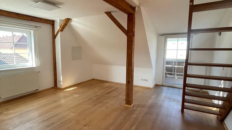 Heller Dachgeschoss-Wohnbereich mit sichtbaren Holzbalken, Holztreppe und natürlichem Lichteinfall durch Fenster.