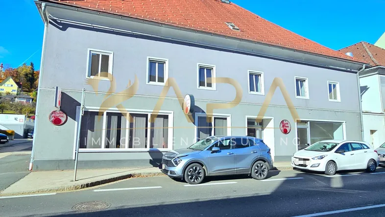 Graues Gebäude mit rotem Ziegeldach und geparkten Autos davor unter blauem Himmel.