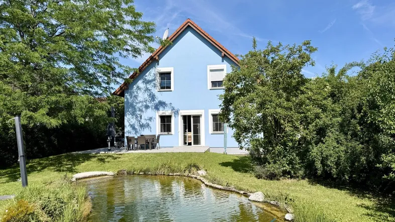 Blaues Haus mit rotem Dach, umgeben von einem grünen Garten und einem natürlichen Teich.