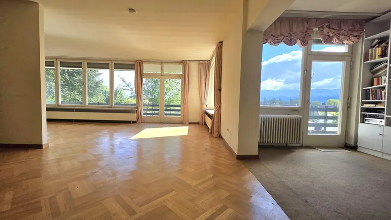 Geräumiges Wohnzimmer mit Fischgrätparkett, großen Fenstern und weitem Blick auf die Landschaft und Berge.