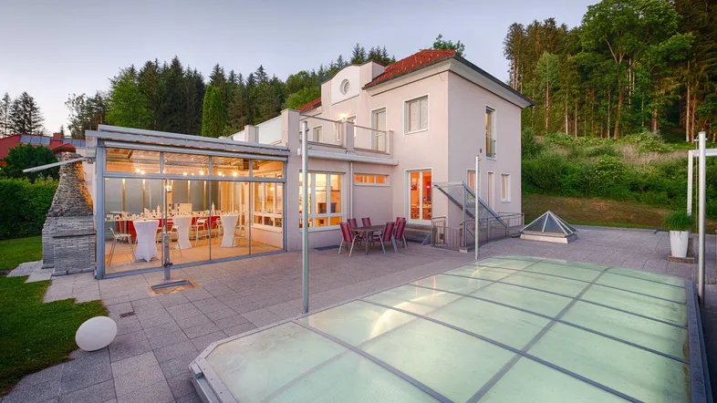 Hausfassade mit Wintergarten, Terrasse und überdachtem Pool in grüner Umgebung.