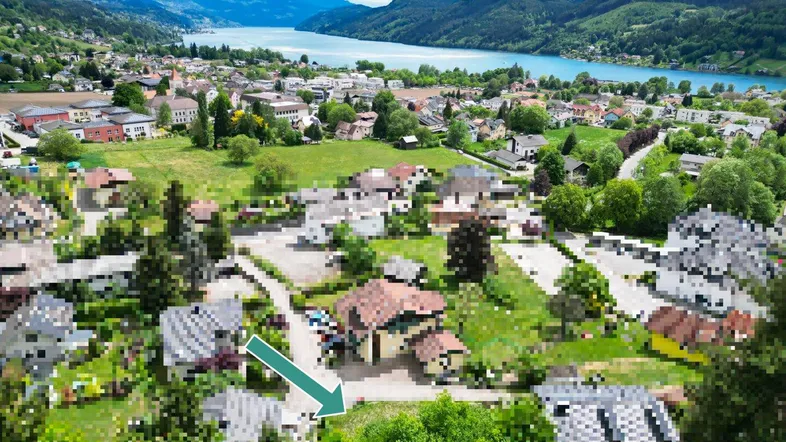 Luftaufnahme eines Dorfes am Seeufer mit Bergen im Hintergrund und einem markierten Grundstück.