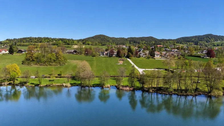 Panoramablick auf den See, grüne Hügel und eine idyllische Siedlung unter blauem Himmel.