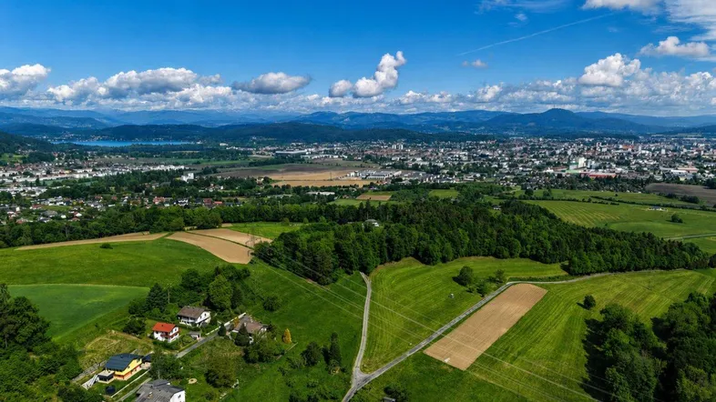 Luftaufnahme einer weiten Landschaft mit Feldern, Wäldern, einer Stadt und Bergen im Hintergrund unter blauem Himmel.