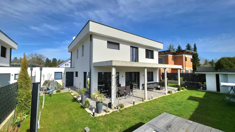Modernes Einfamilienhaus mit großer Terrasse und gepflegtem Garten unter blauem Himmel.