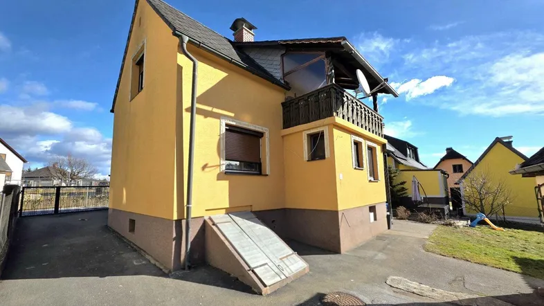 Gelbes Einfamilienhaus mit braunem Sockel, einem Balkon und Zugang zum Keller.