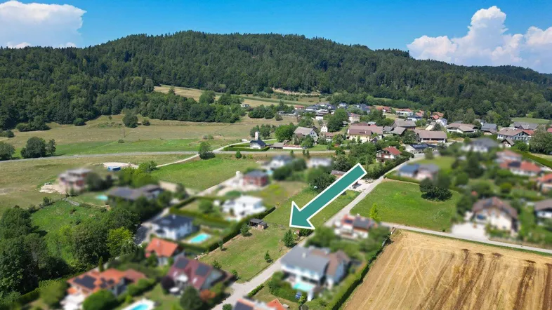 Luftaufnahme einer Wohnsiedlung mit Häusern, grünen Feldern und einem Wald im Hintergrund, mit Bergblick.