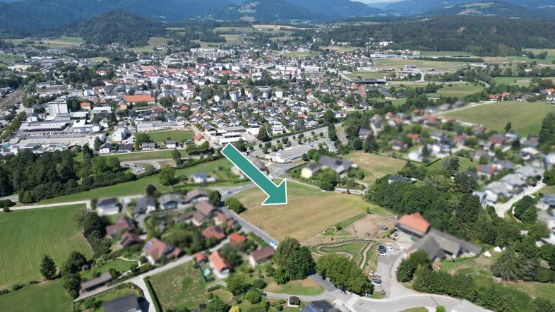 Luftaufnahme einer ländlichen Siedlung mit einem freien Grundstück, umgeben von Häusern und Bergen.