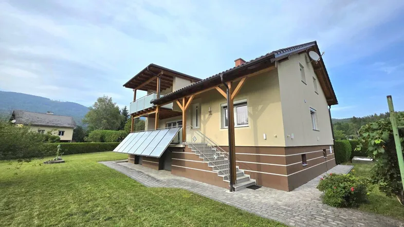 Hausfassade mit Garten, Balkon und Solarpaneelen unter blauem Himmel.