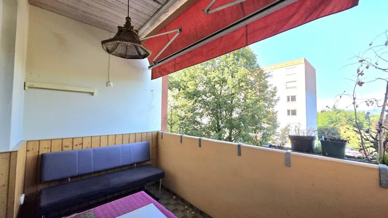 Gemütlicher Balkon mit Sitzgelegenheit, roter Markise und Blick ins Grüne.