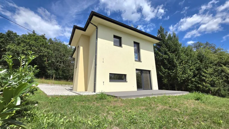 Modernes Einfamilienhaus mit heller Fassade, umgeben von Grünflächen und Bäumen unter blauem Himmel.