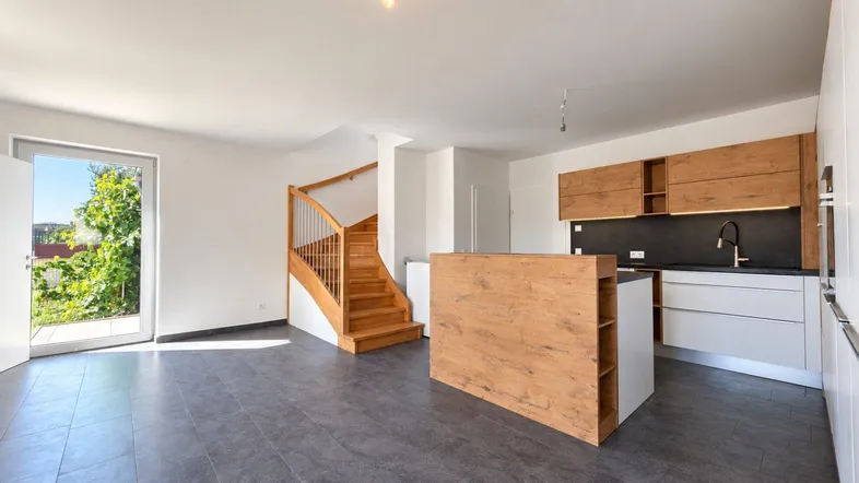 Heller offener Wohn- und Essbereich mit moderner Küche, Fliesenboden und Holztreppe zum Obergeschoss.