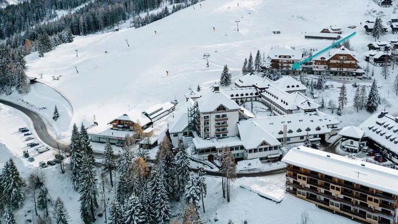 Luftaufnahme eines großen Gebäudekomplexes in einer verschneiten Berglandschaft mit Skipisten und umliegenden Wäldern.
