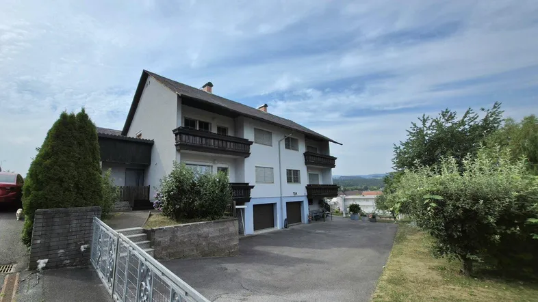 Mehrfamilienhaus mit Balkonen, Garagen und einer Zufahrt in ländlicher Umgebung unter blauem Himmel.