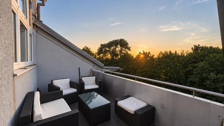Gemütlicher Balkon mit Rattanmöbeln und Blick auf den Sonnenuntergang über den Bäumen.