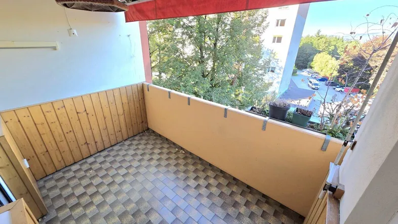 Geräumiger Balkon mit Holzwänden, Fliesenboden und roter Markise, bietet einen Blick ins Grüne.
