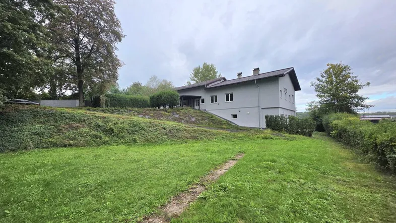 Graues Haus mit Garten und Bäumen unter bewölktem Himmel, umgeben von viel Grün.