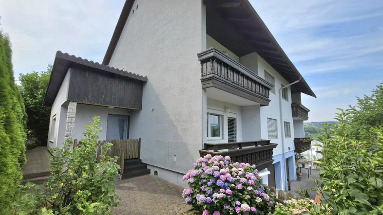 Mehrfamilienhaus mit traditionellen Holzbalkonen und gepflegter Außenanlage mit blühenden Hortensien.