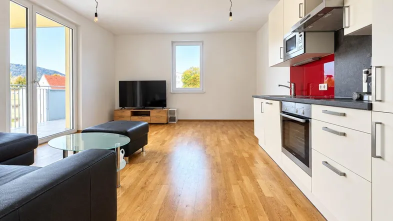 Heller offener Wohnbereich mit integrierter Küche, Parkettboden und Zugang zum Balkon.