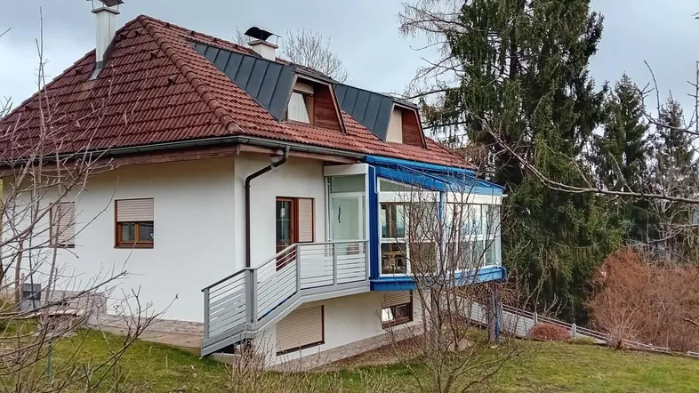 Freistehendes Haus mit rotem Ziegeldach und einem verglasten Wintergarten, umgeben von Bäumen.