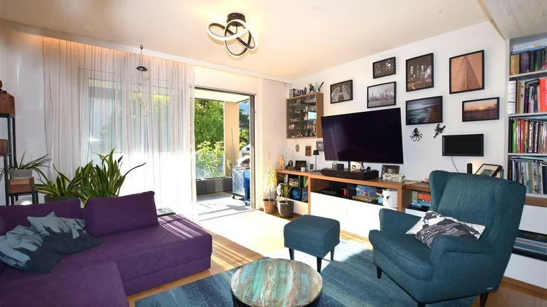 Helles Wohnzimmer mit lila Sofa, blauem Teppich und Zugang zum sonnigen Balkon.