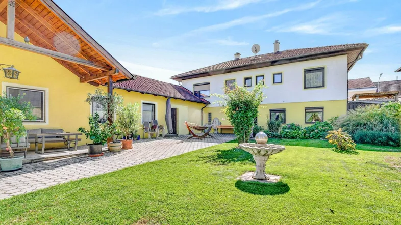 Großer Gartenbereich mit gepflasterter Terrasse, Rasenfläche und dekorativem Brunnen zwischen zwei Gebäuden.