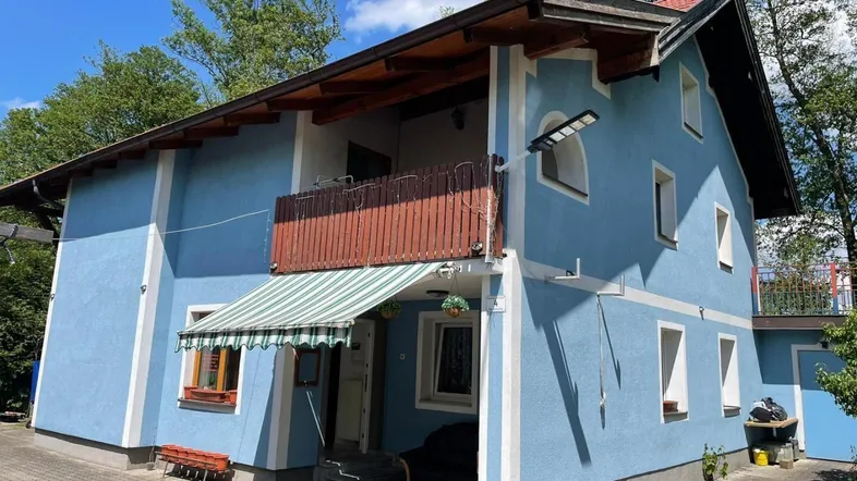 Blaues Wohnhaus mit Holzbalkon, Markise und gepflastertem Vorplatz unter blauem Himmel.