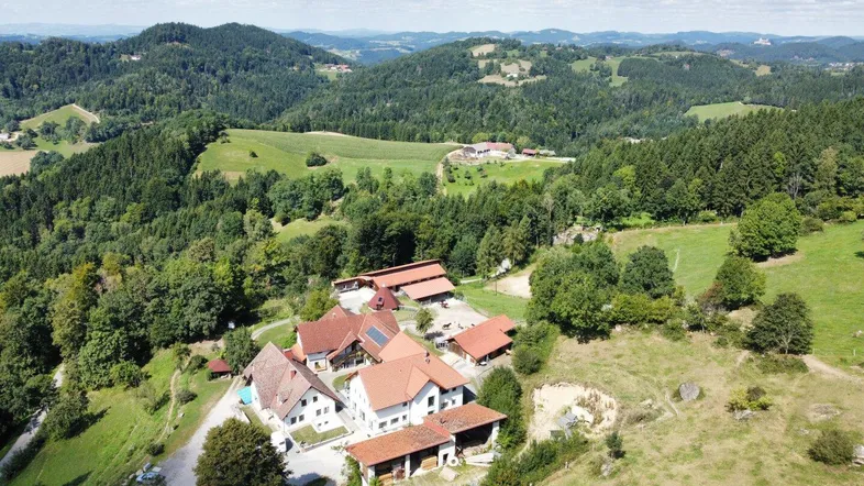 Luftaufnahme eines weitläufigen Anwesens mit mehreren Gebäuden, umgeben von grünen Hügeln und Wäldern.