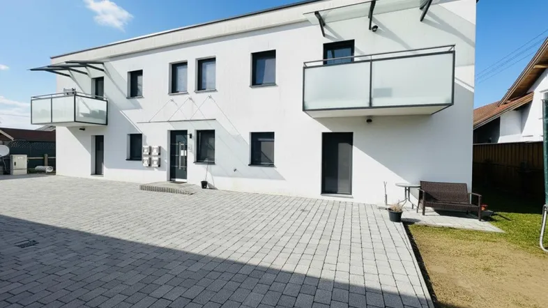 Moderne weiße Fassade eines Mehrfamilienhauses mit Balkonen und gepflastertem Vorplatz unter blauem Himmel.