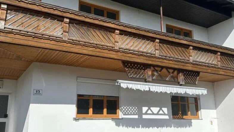 Fassade eines traditionellen Hauses mit weißem Putz und einem langen Holzbalkon im Obergeschoss.