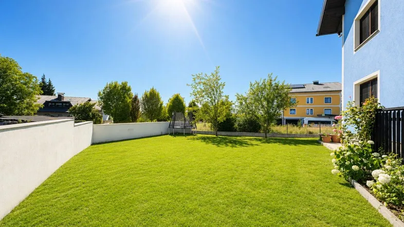 Großer, gepflegter Rasenbereich mit Trampolin und sonniger Ausrichtung neben dem blauen Haus.