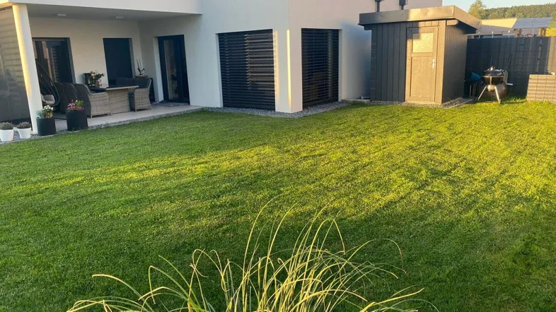 Modernes Haus mit großem, gepflegtem Rasen, einer möblierten Terrasse und einem Gartenschuppen.
