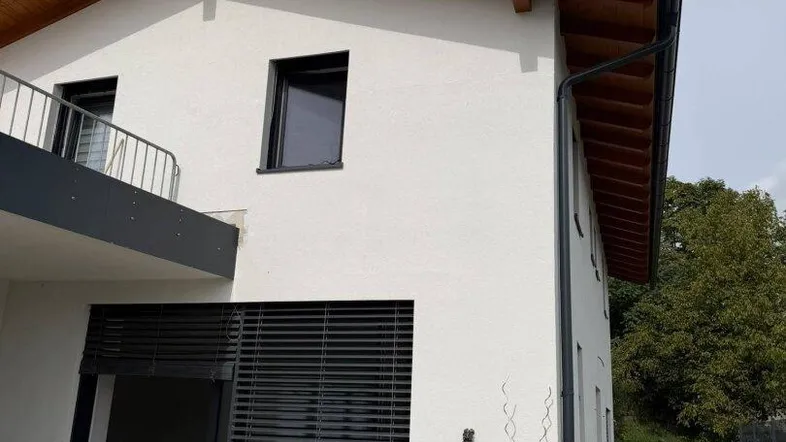 Moderne Hausfassade mit weißem Putz, dunklen Fenstern und Holzdachüberstand.