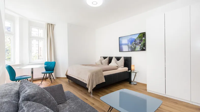 Helles Wohnzimmer mit Schlafbereich, Sofa, Esstisch und großem Flachbildfernseher.