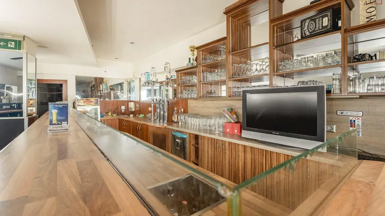 Blick auf die Theke einer Bar mit Holzoberfläche, gefüllten Regalen und einem Fernseher.