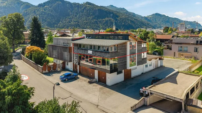 Luftaufnahme eines modernen Mehrzweckgebäudes mit Holzfassade und großen Fenstern vor einer Bergkulisse.